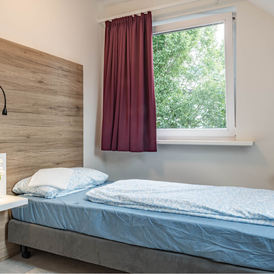 Single bed in a small room with wooden headboard, window with curtain, and a wall lamp.