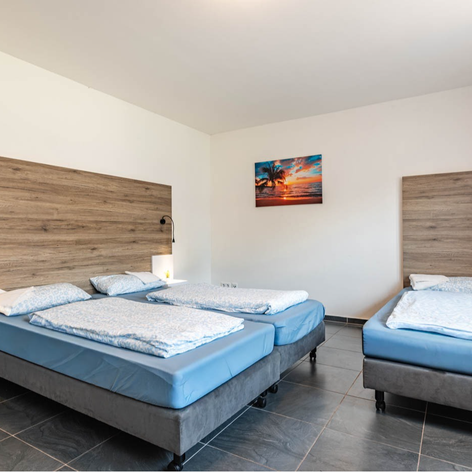 Bedroom with three beds: two twin beds together, a single bed, light walls, wooden headboards, and a sunset painting.