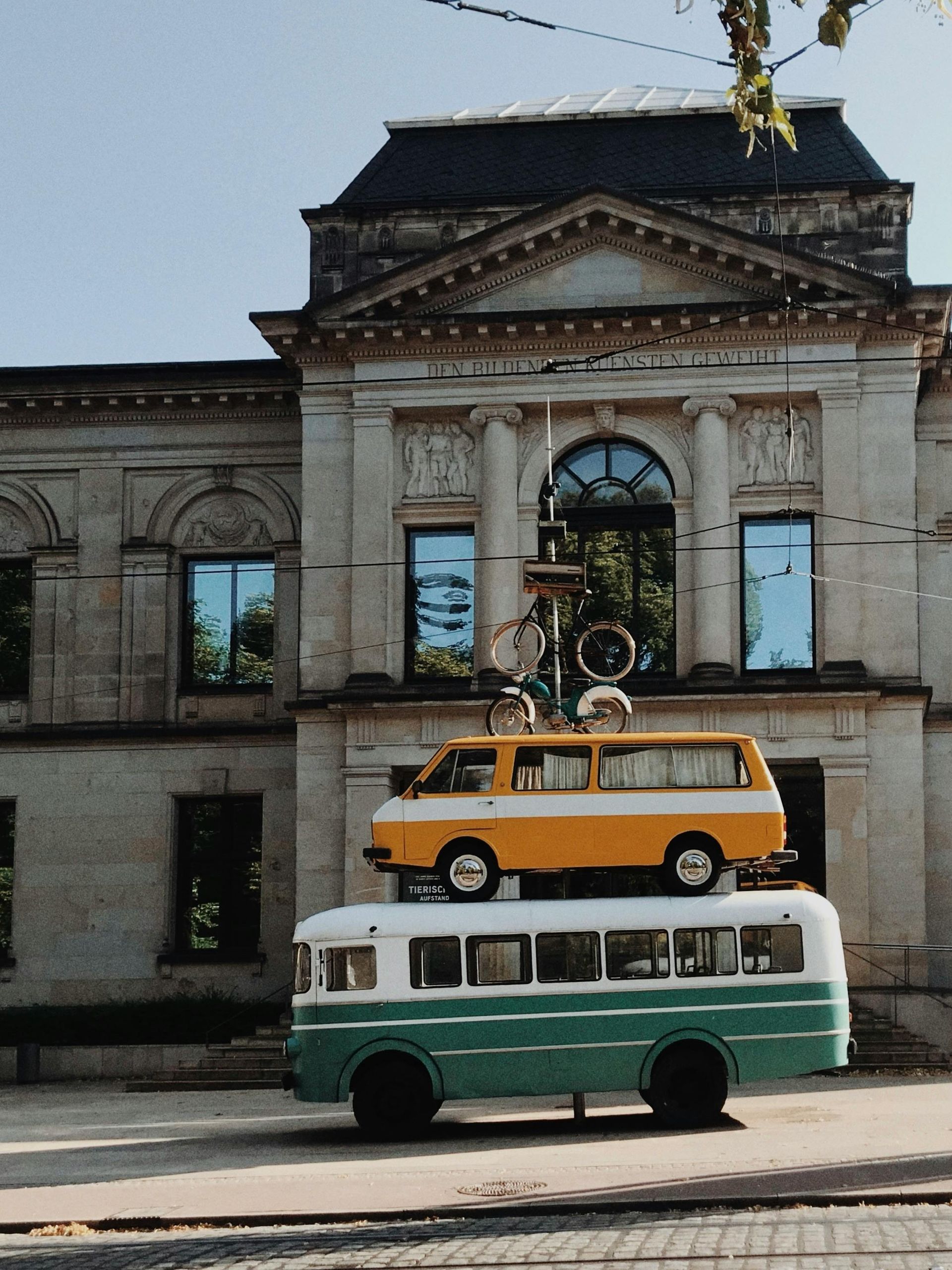 Green and white bus stacked with a yellow van and a bike on top, in front of a stone building with blue-tinted windows.