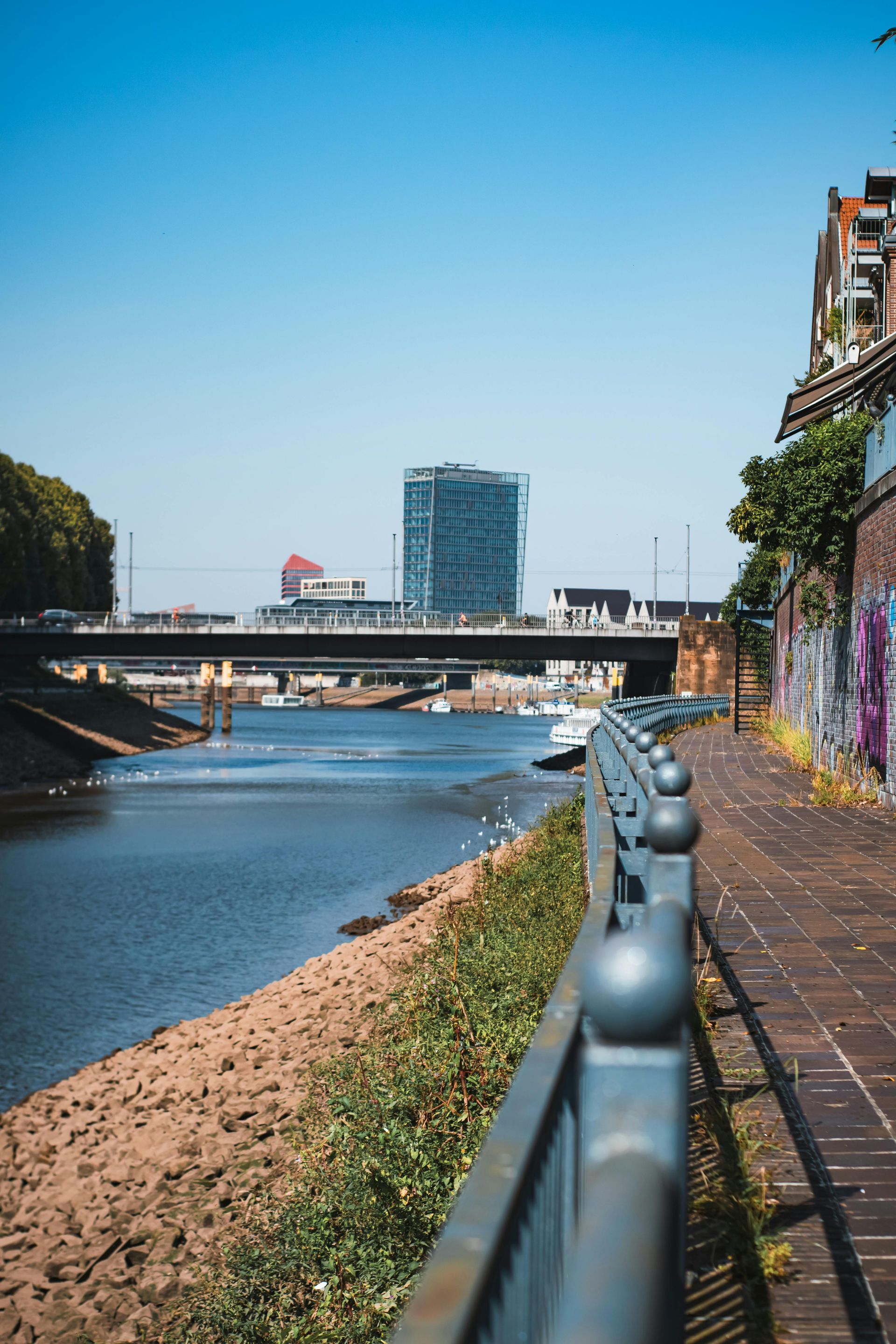 Waterfront scene with a river, bridge, and tall building under a clear blue sky.