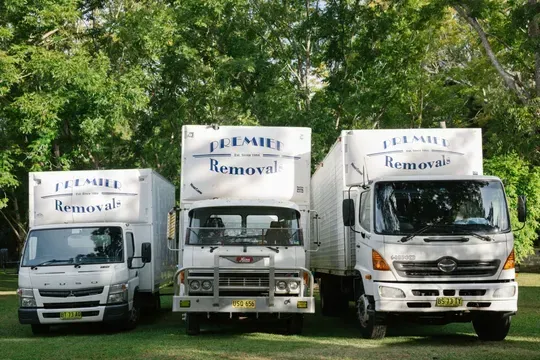 Three White Moving Trucks Parked on Grass in Front of Trees — Premier Removals & Storage In Boambee, NSW
