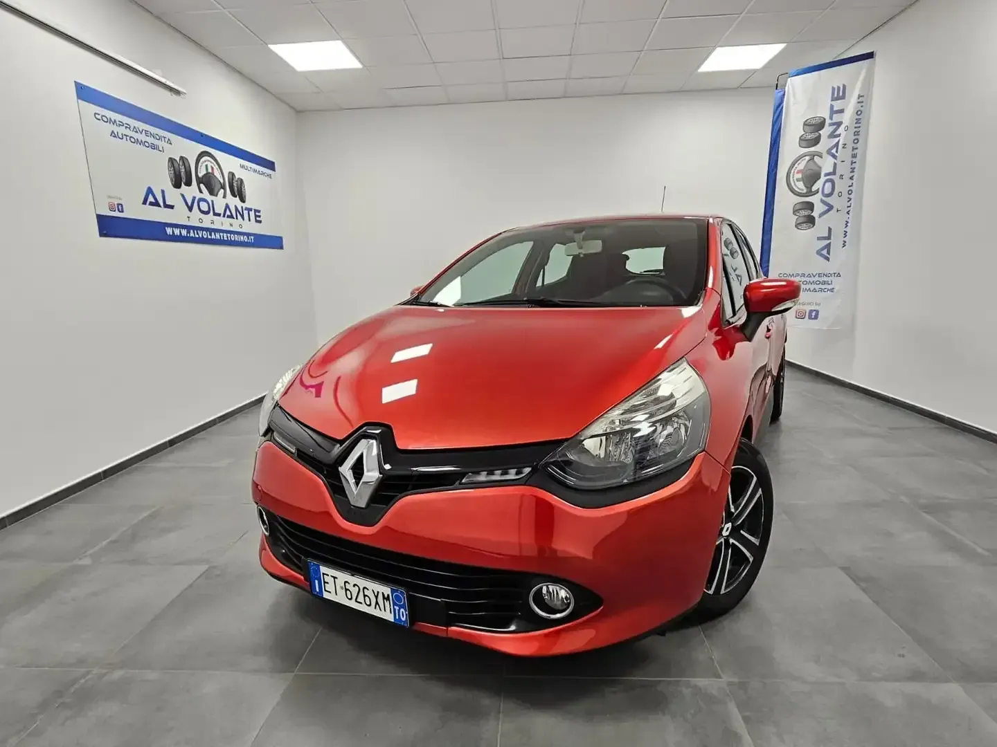 Una Renault Clio hatchback rossa parcheggiata nello showroom luminoso e moderno di una concessionaria, con pareti bianche e pavimento piastrellato.