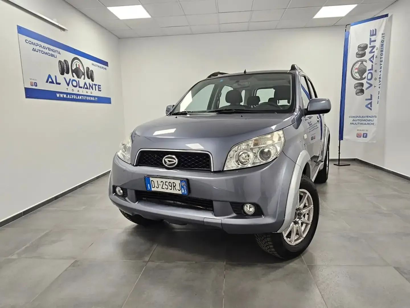 Una Daihatsu Terios grigia parcheggiata in uno showroom interno luminoso con pareti bianche e striscioni pubblicitari.