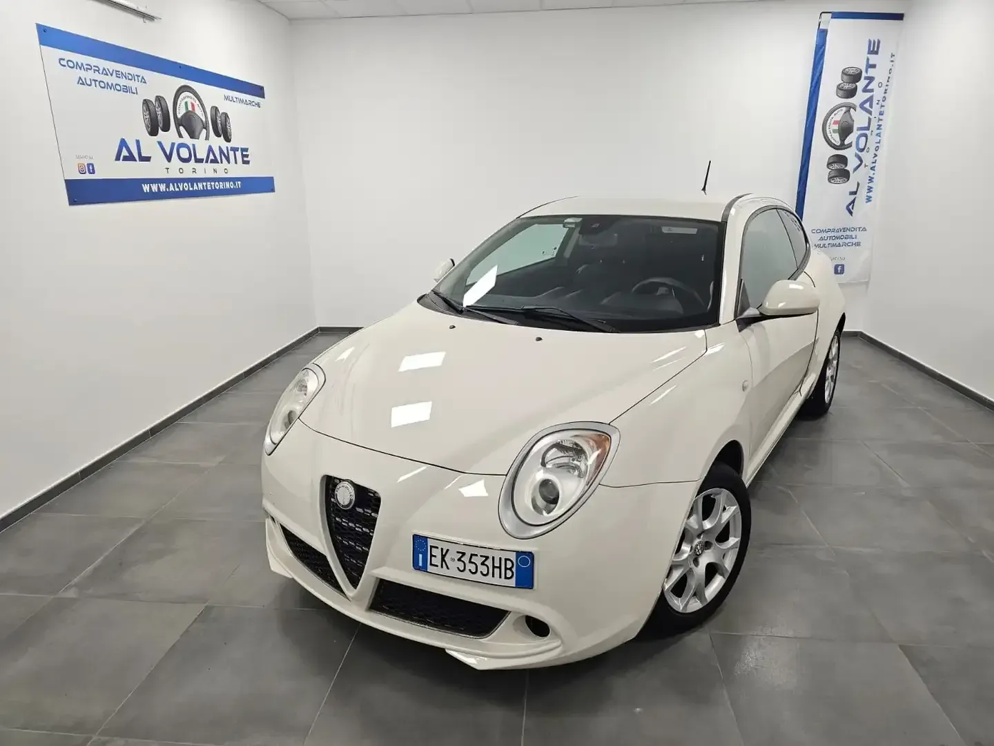 Un'Alfa Romeo MiTo bianca parcheggiata in uno showroom ben illuminato, con poster del marchio alle pareti.