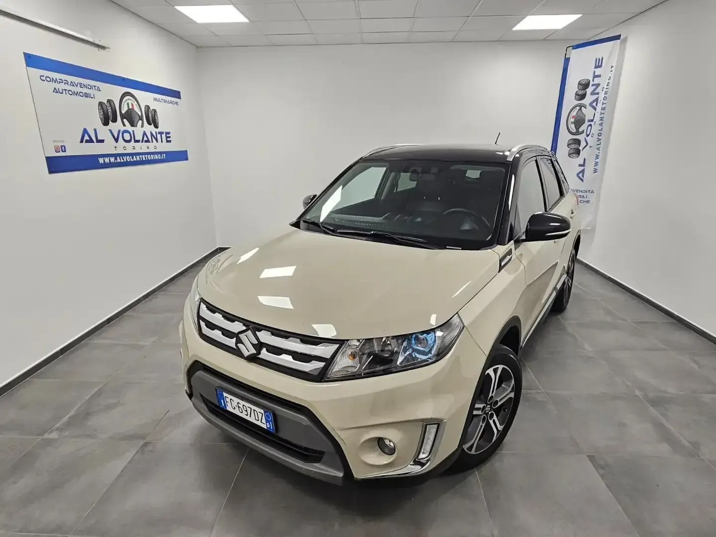 Un SUV Suzuki Vitara beige e nero parcheggiato all'interno di uno showroom luminoso con pareti bianche e un'insegna con il logo sulla parete.