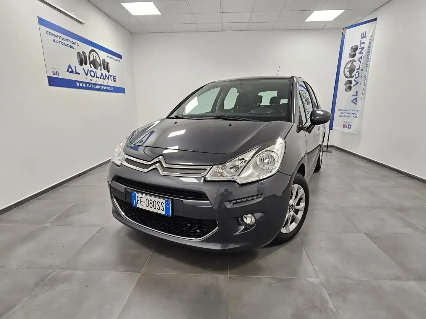 Una Citroën C3 grigio scuro parcheggiata in uno showroom luminoso con pareti bianche e uno striscione con il logo sulla parete.