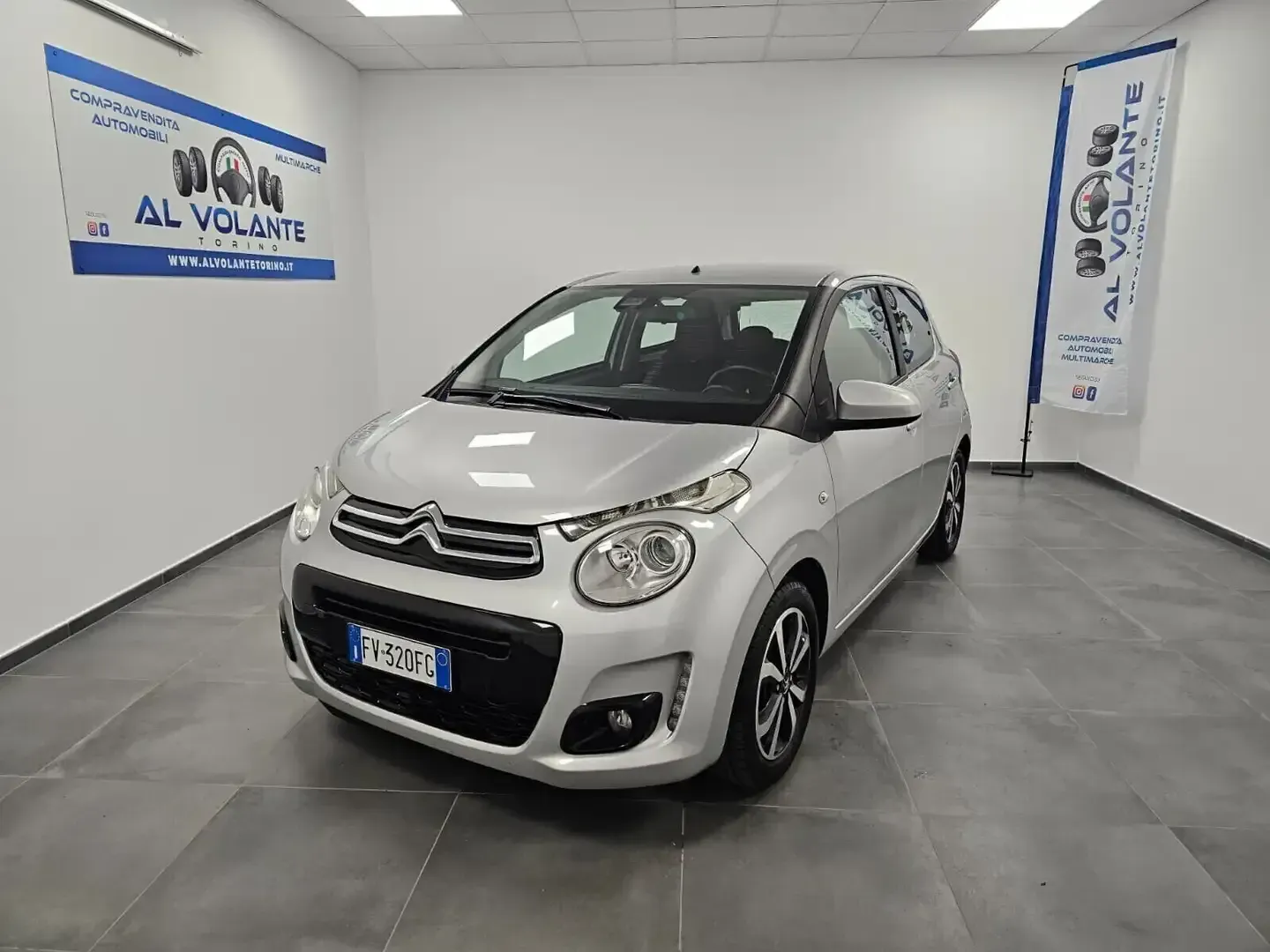 Una Citroën C1 compatta color argento parcheggiata in uno showroom luminoso con pareti bianche e striscioni del marchio sulle pareti laterali.