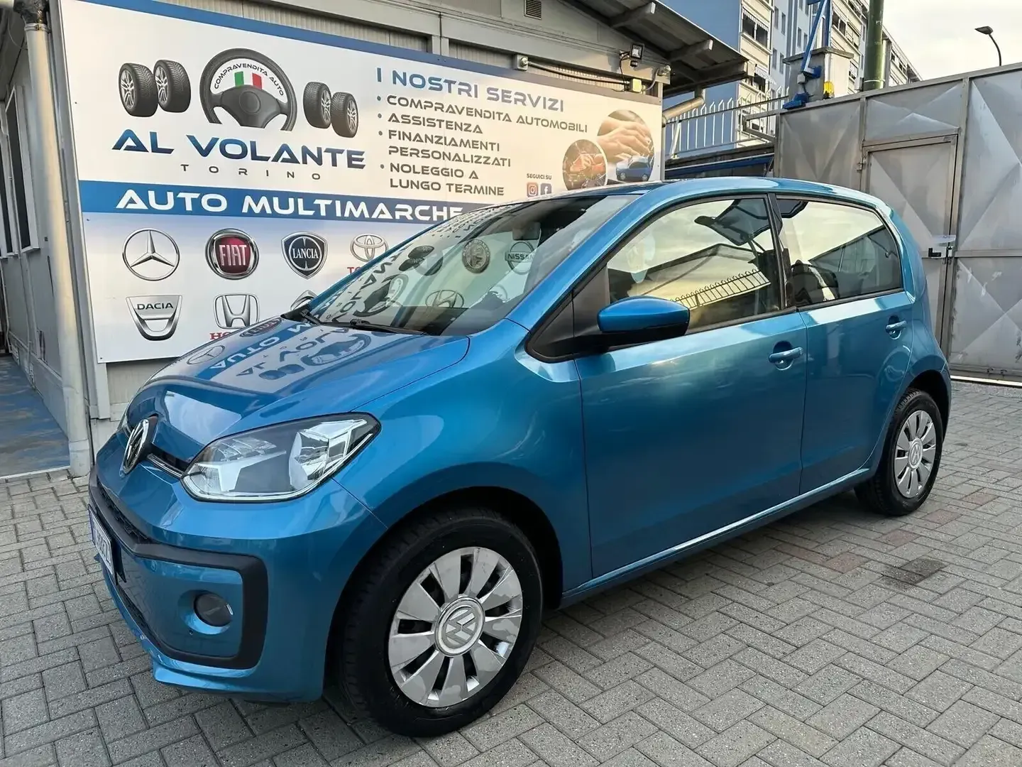 Una Volkswagen Up blu parcheggiata davanti a un'officina meccanica con un cartello che elenca i servizi offerti dall'azienda.