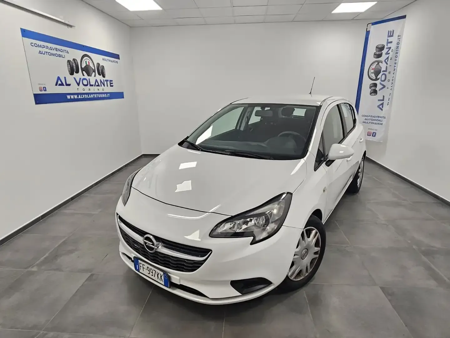 Una Opel Corsa hatchback bianca parcheggiata nello showroom interno di una concessionaria, luminoso e con pareti bianche.