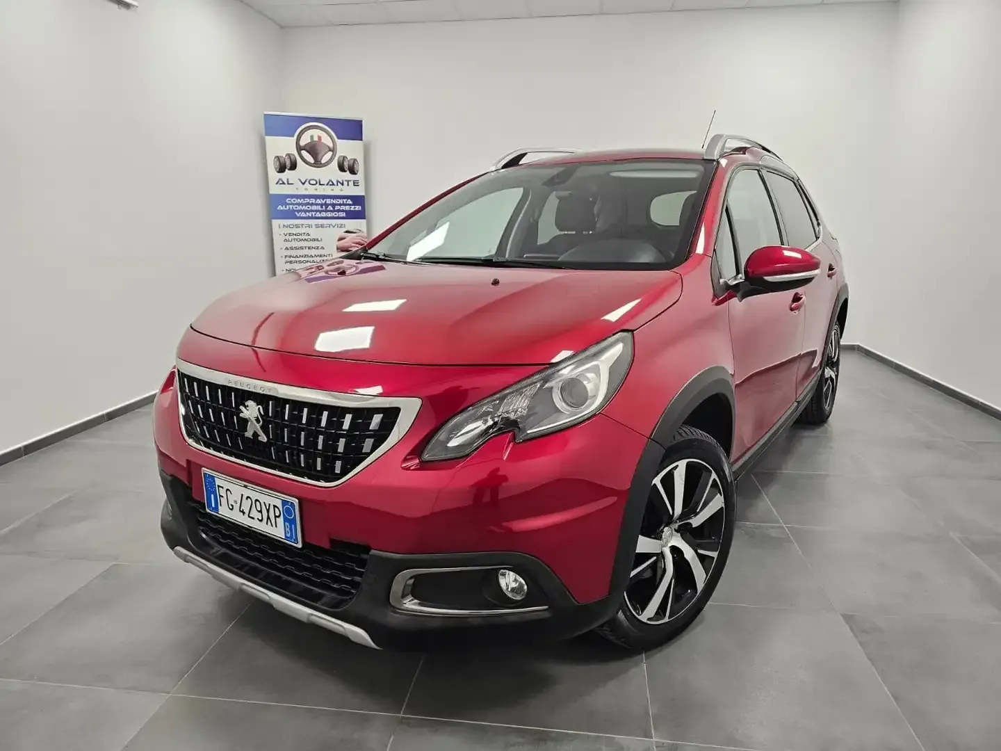 Un SUV Peugeot 2008 rosso parcheggiato in uno showroom luminoso con pareti bianche e uno striscione sullo sfondo.