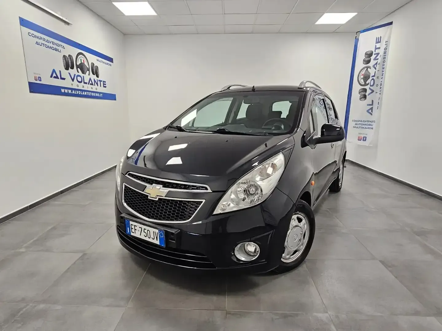 Una Chevrolet Spark nera parcheggiata all'interno di uno showroom di una concessionaria ben illuminato, con pareti bianche e pavimenti piastrellati.