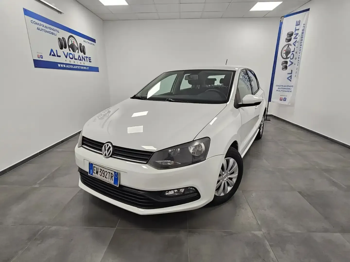 Una Volkswagen Polo bianca parcheggiata in un luminoso showroom al coperto di una concessionaria d'auto, con pavimento piastrellato di grigio.