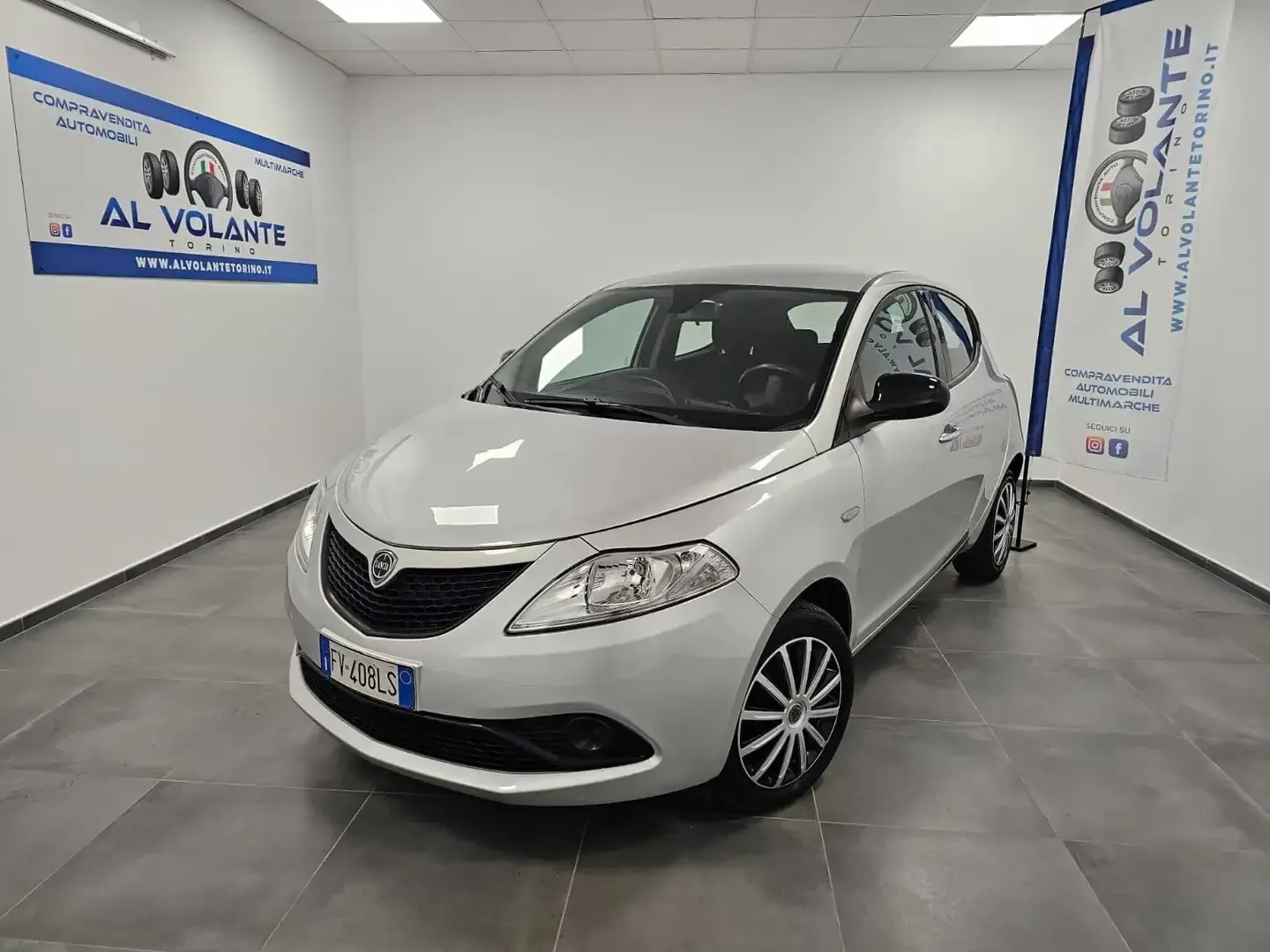 Una Lancia Ypsilon color argento parcheggiata in uno showroom ben illuminato, con il logo della concessionaria sulle pareti bianche.