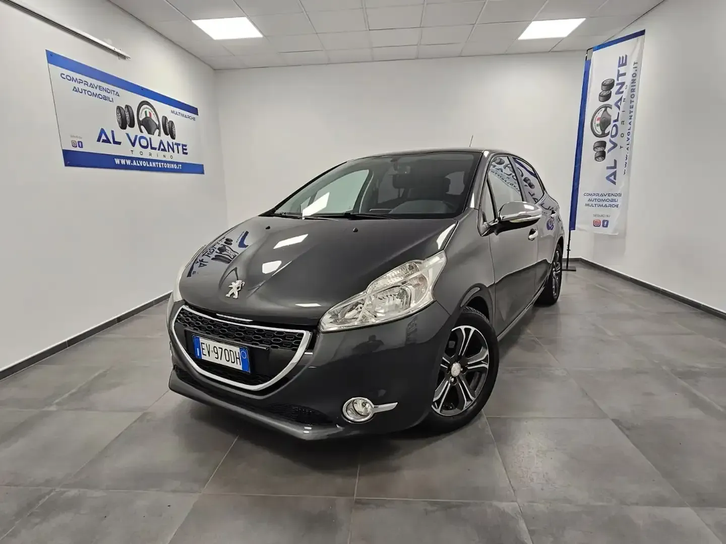 Una Peugeot 208 grigio scuro parcheggiata in uno showroom luminoso con pareti bianche e un'insegna con il logo sulla parete.