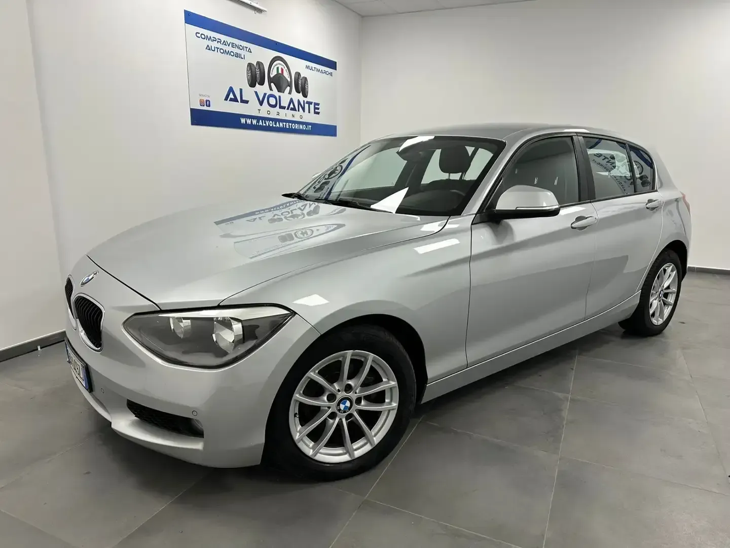 Una BMW Serie 1 hatchback color argento parcheggiata in un luminoso showroom al coperto con pavimento piastrellato e un cartello sulla parete.