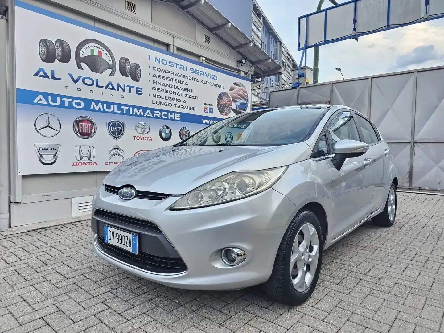 Una Ford Fiesta color argento parcheggiata davanti a una concessionaria con i loghi del marchio sulle pareti.
