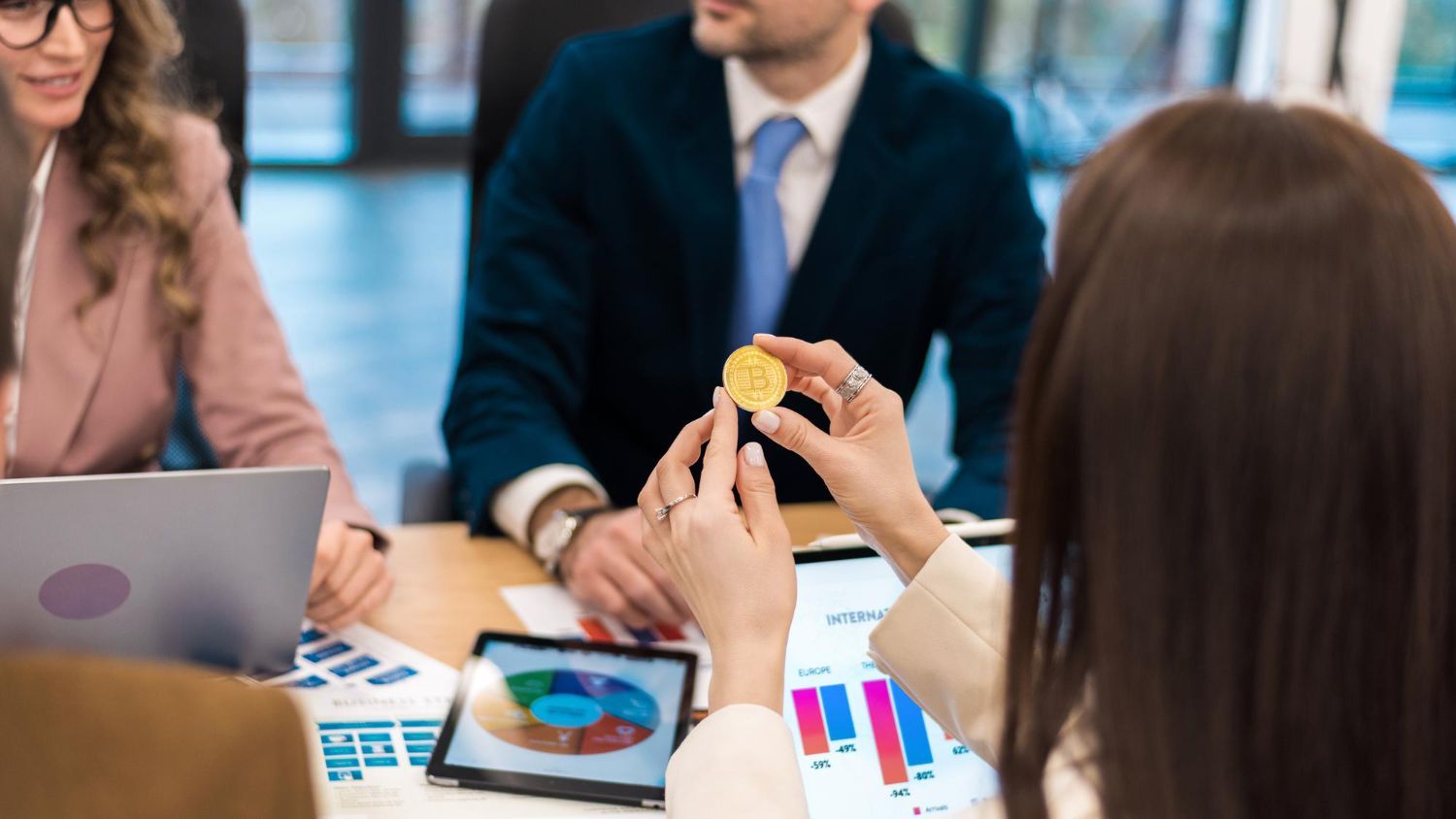 Reunião de negócios com pessoas ao redor de uma mesa analisando gráficos e um tablet; uma pessoa segura um objeto amarelo.