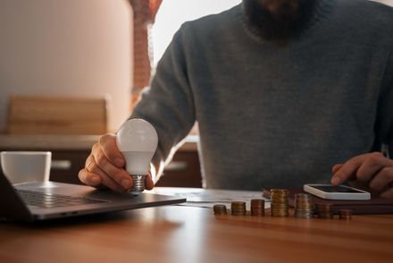 Pessoa contando moedas ao lado de um laptop e um smartphone em uma mesa de madeira, com uma lâmpada na mão.