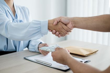 Duas pessoas apertam as mãos sobre uma mesa com papéis e uma prancheta em um escritório bem iluminado.