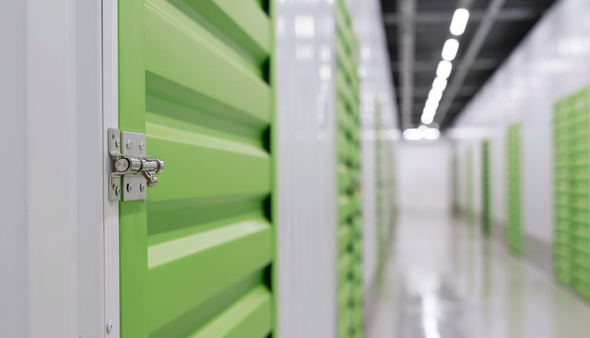 Green storage unit door with silver latch in a brightly lit hallway of units.