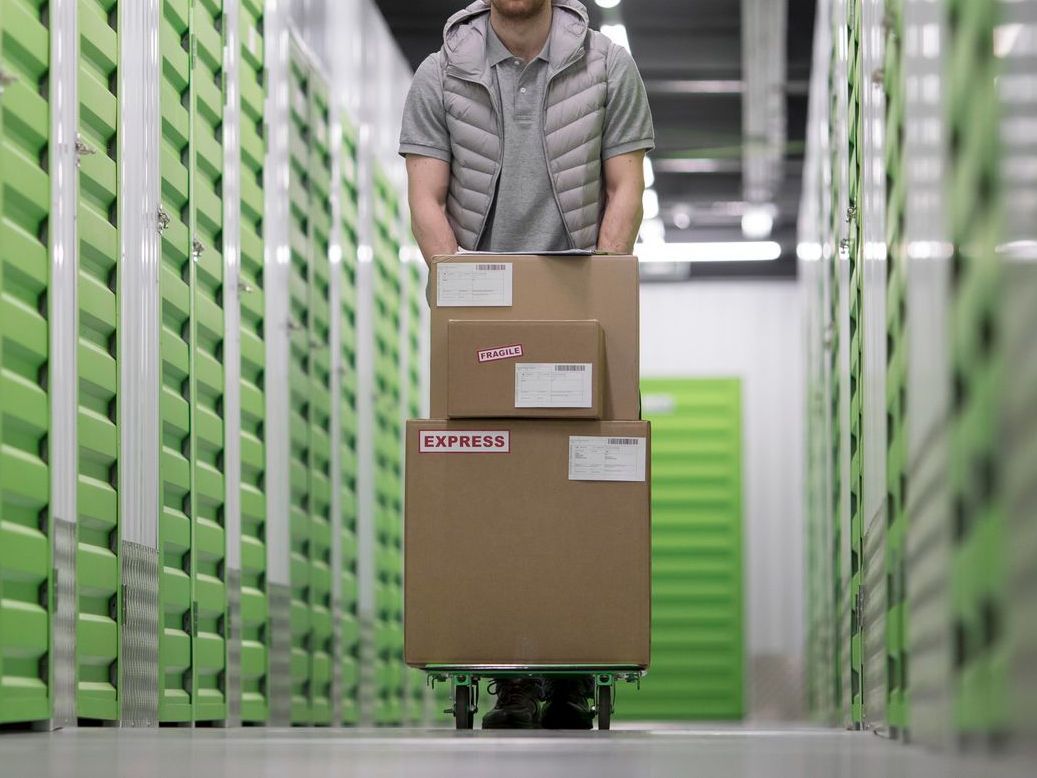 Person pushing boxes on a cart down a storage unit hallway. Green and white units.