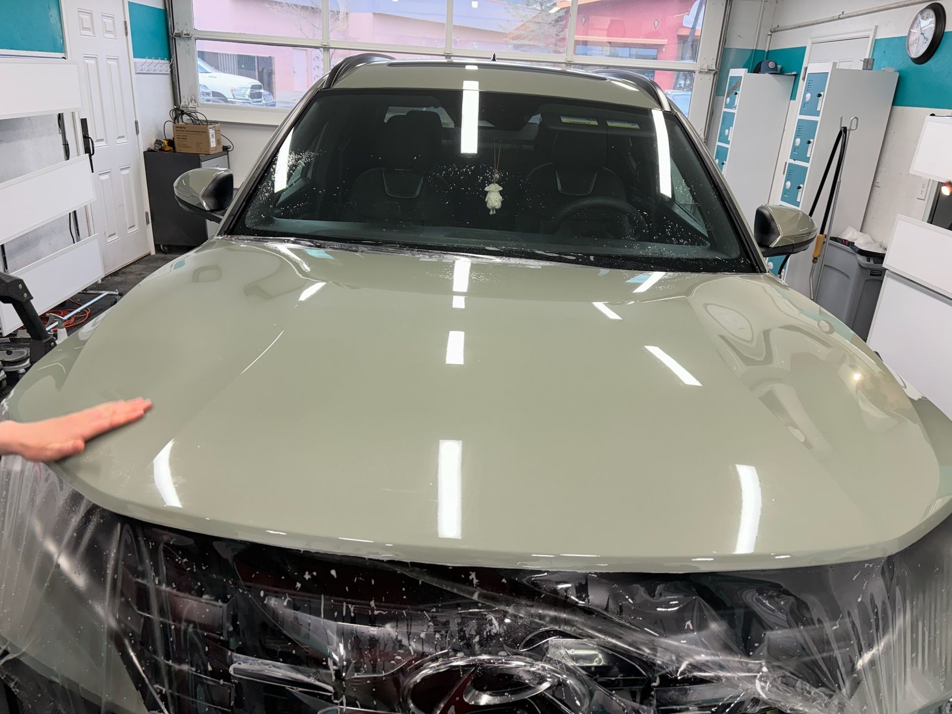 Car hood wrapped in protective film, light green. Hand smoothing the film. Workshop setting.