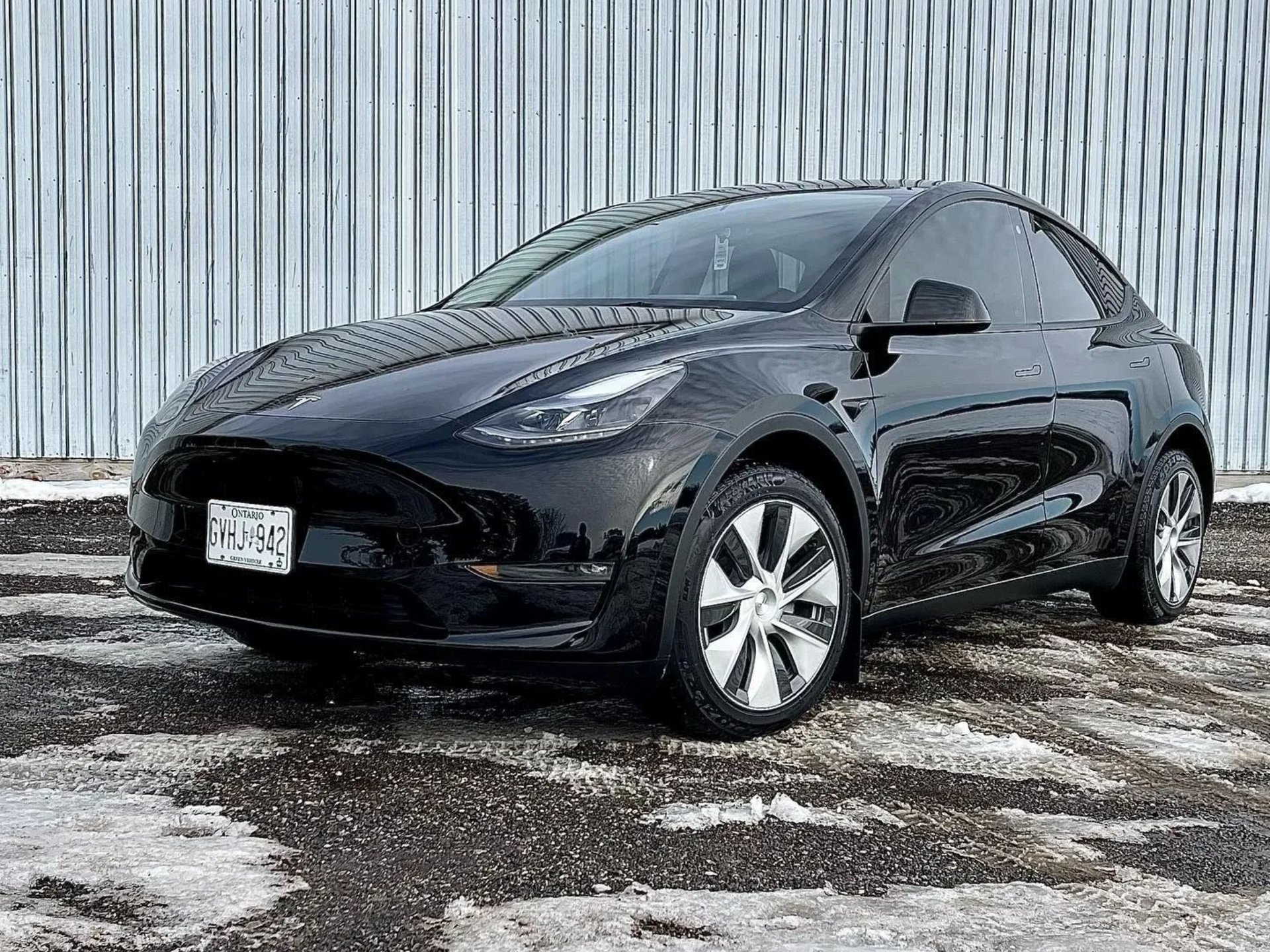 Black Tesla Model Y parked on a snowy, cracked surface, in front of a gray metal wall.