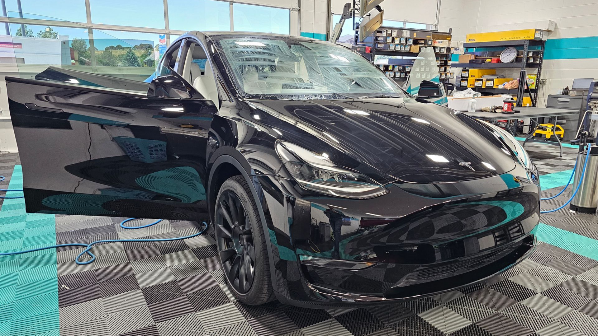 Black Tesla Model Y with open door, parked inside a detailing shop with a tiled floor.