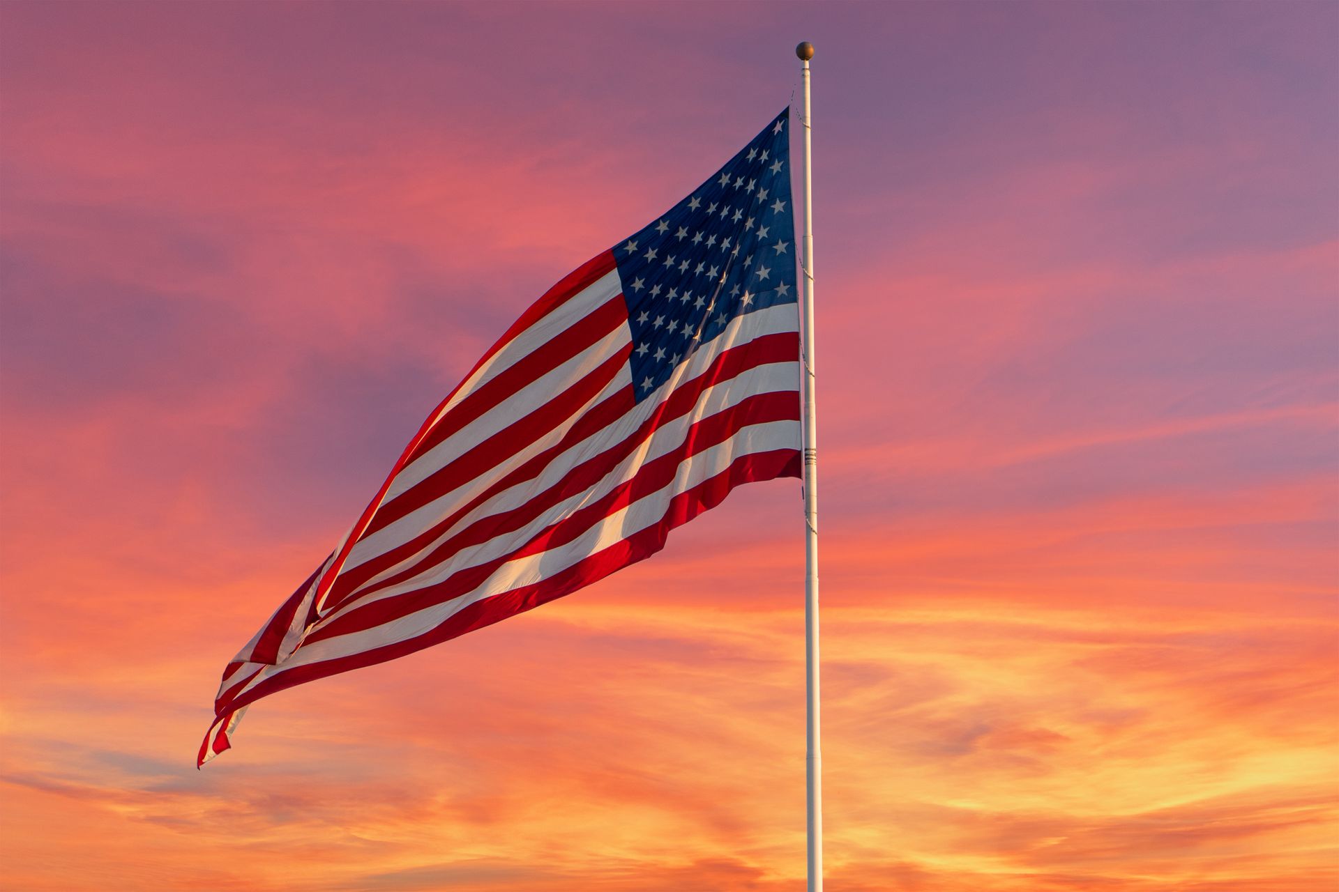 The American flag waves on a pole against a vibrant, orange and pink sunset sky.