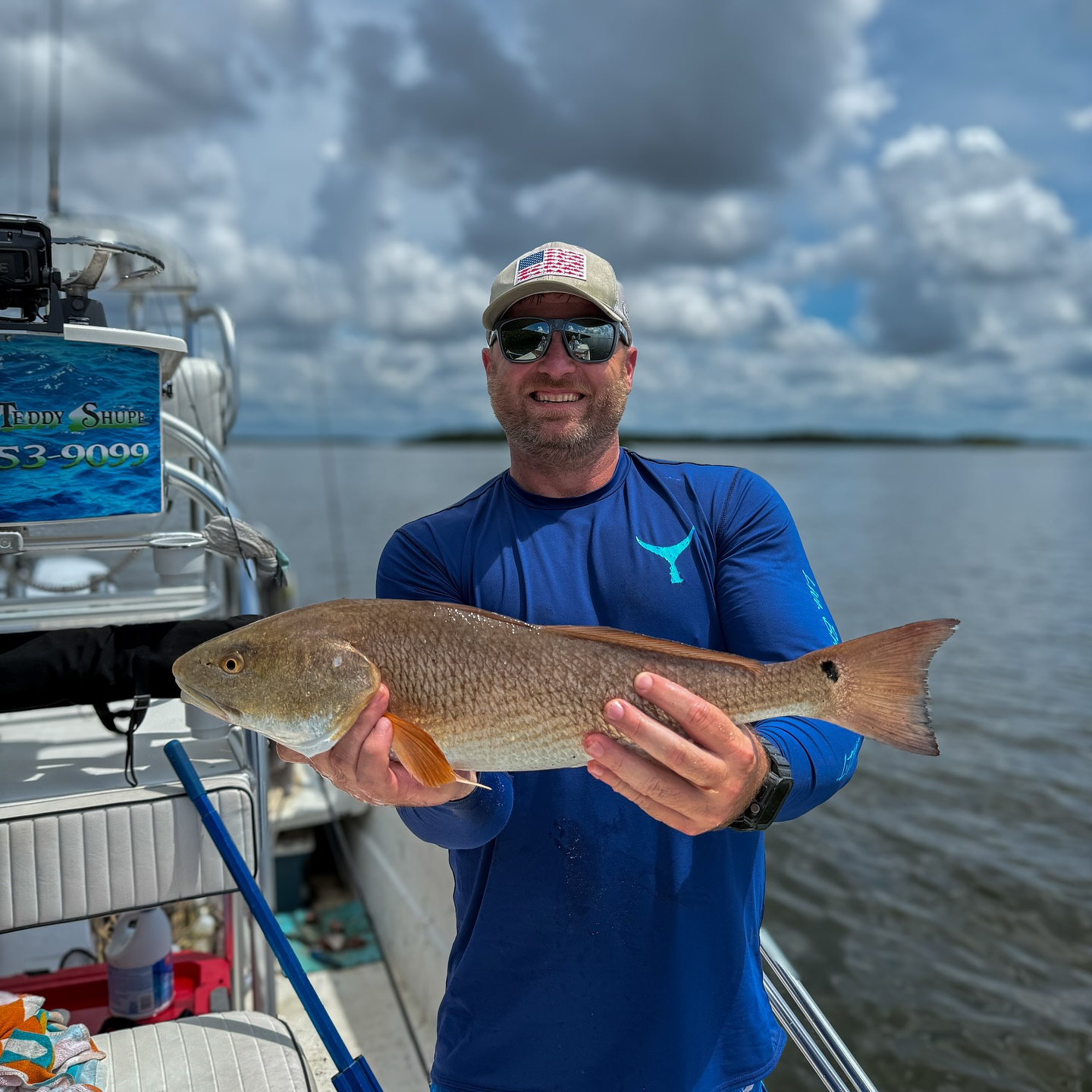 Man on boat holds a large reddish-brown fish, smiling. Blue skies and water in the background.