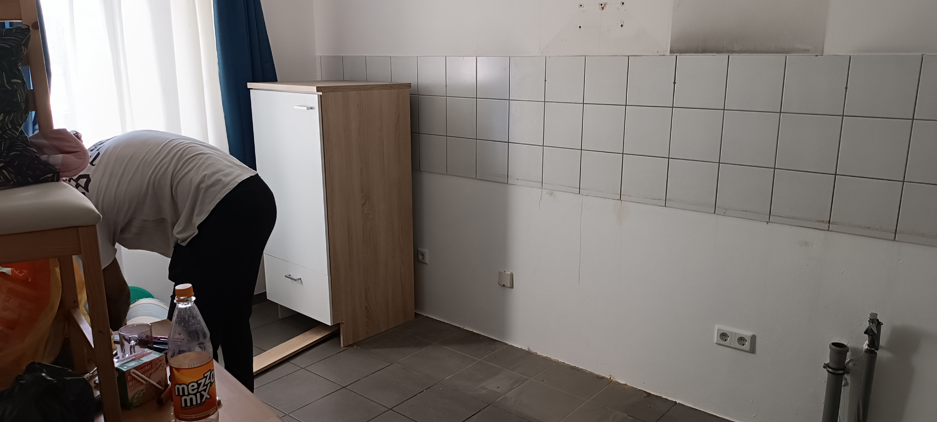Person bückt sich beim Zusammenbauen von Möbeln in einem Raum mit weiß gefliesten Wänden und einem Fenster mit Vorhängen.