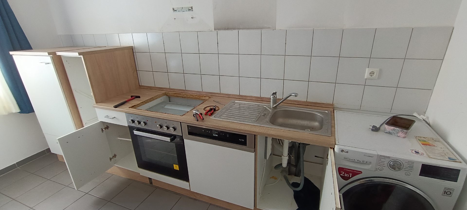 Kleine Küche mit weißen Schränken, Geräten und Fliesenspiegel. Links ein Fenster mit blauen Vorhängen.