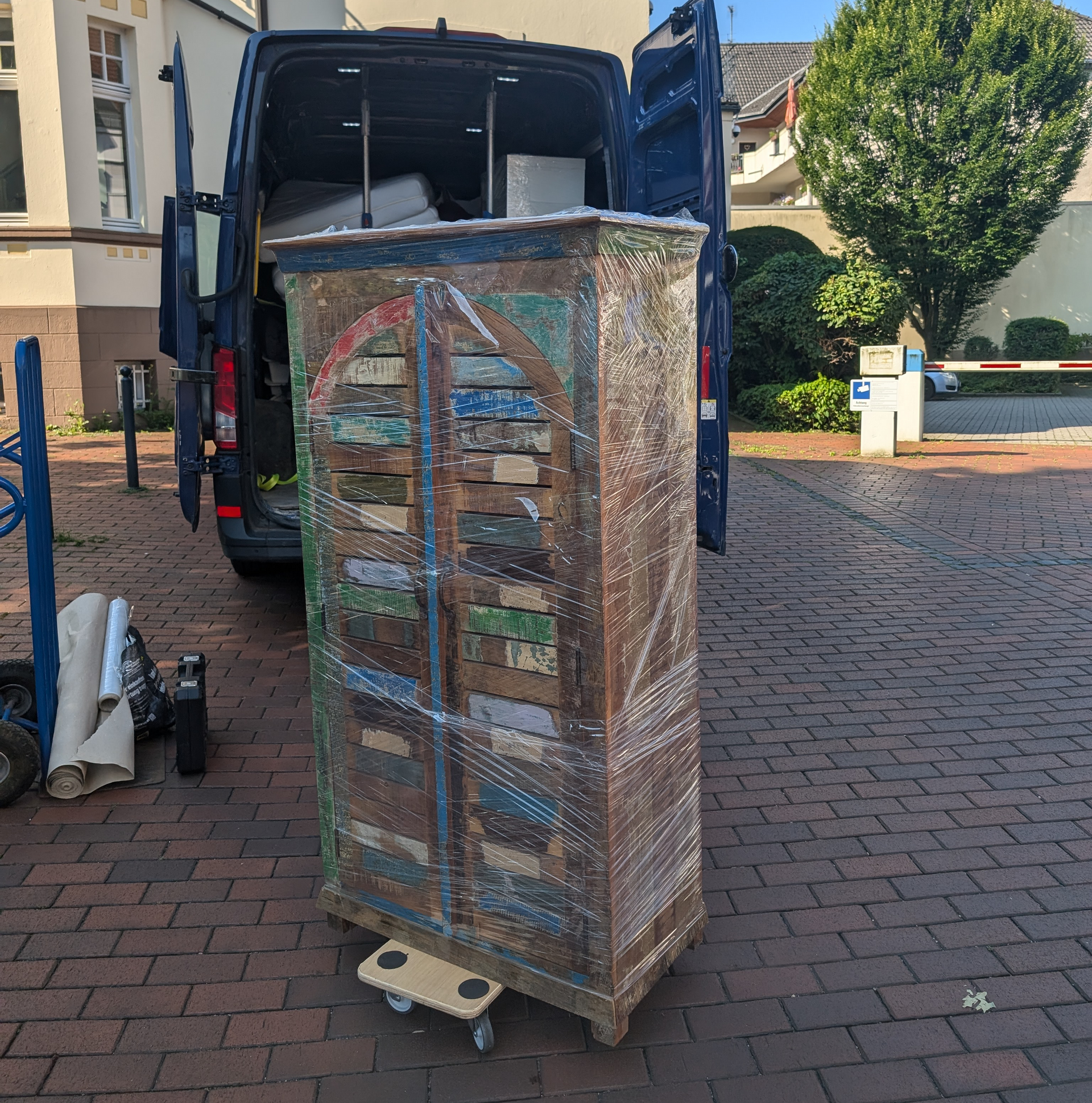 Ein großer antiker Schrank, der in Plastik eingewickelt ist und auf einem Rollwagen aus einem Lieferwagen transportiert wird.
