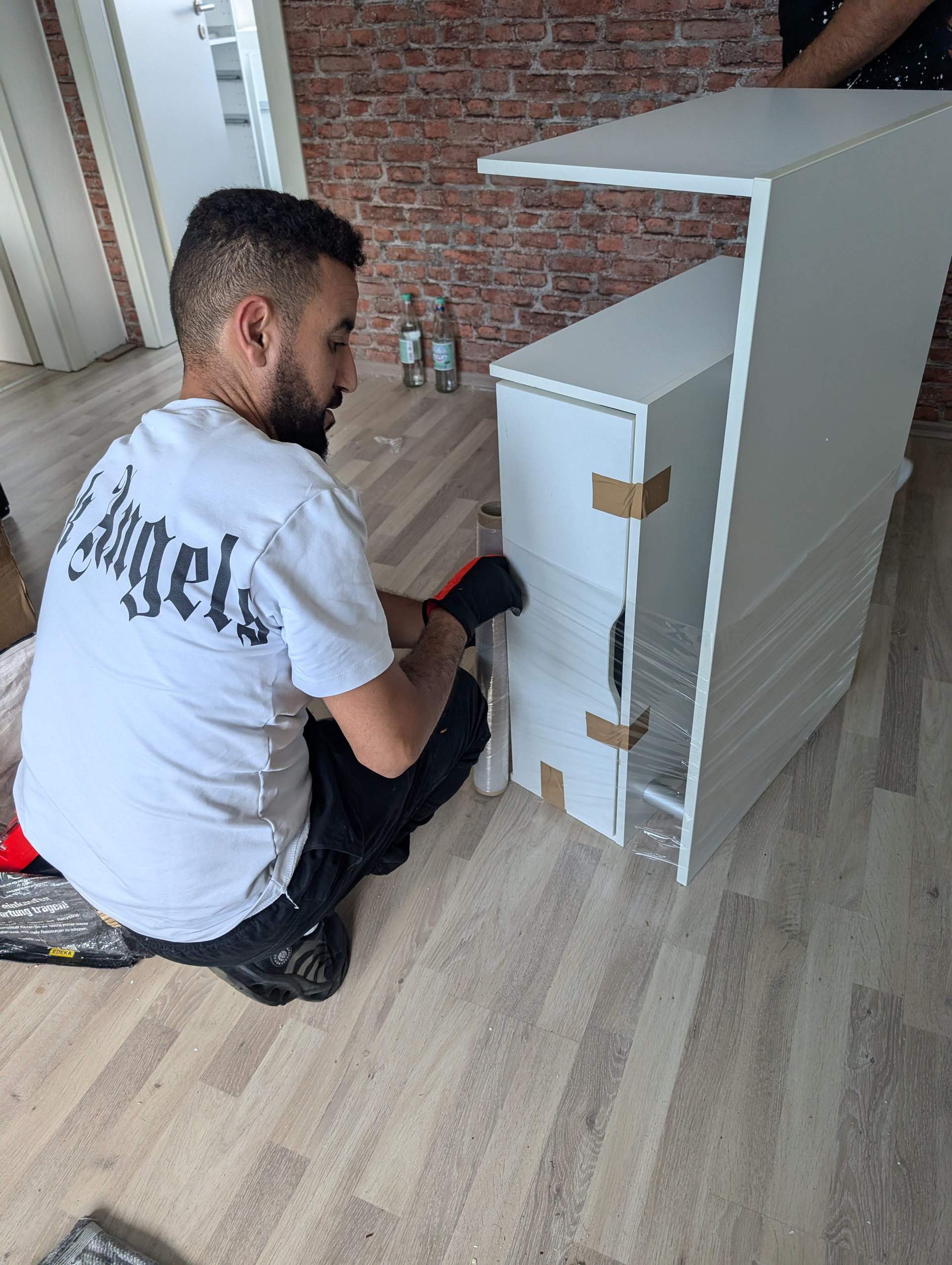 Mann im weißen Hemd baut weiße Möbel neben einer Ziegelwand zusammen.