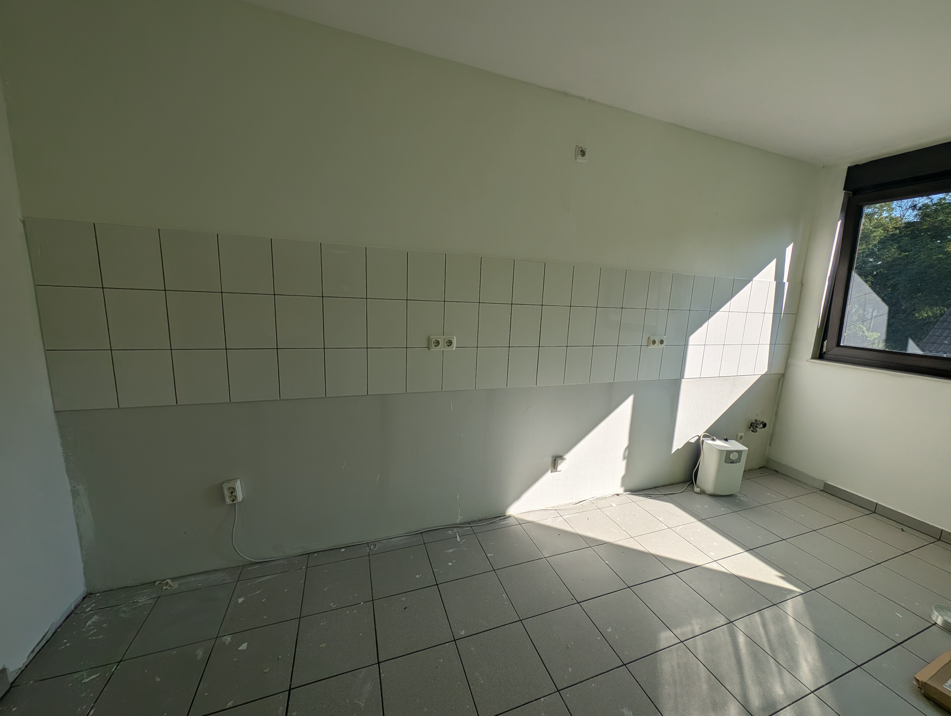 Raum mit teilweise gefliester Wand, Fenster und Boden mit Gittermuster; Sonnenlicht strömt herein.