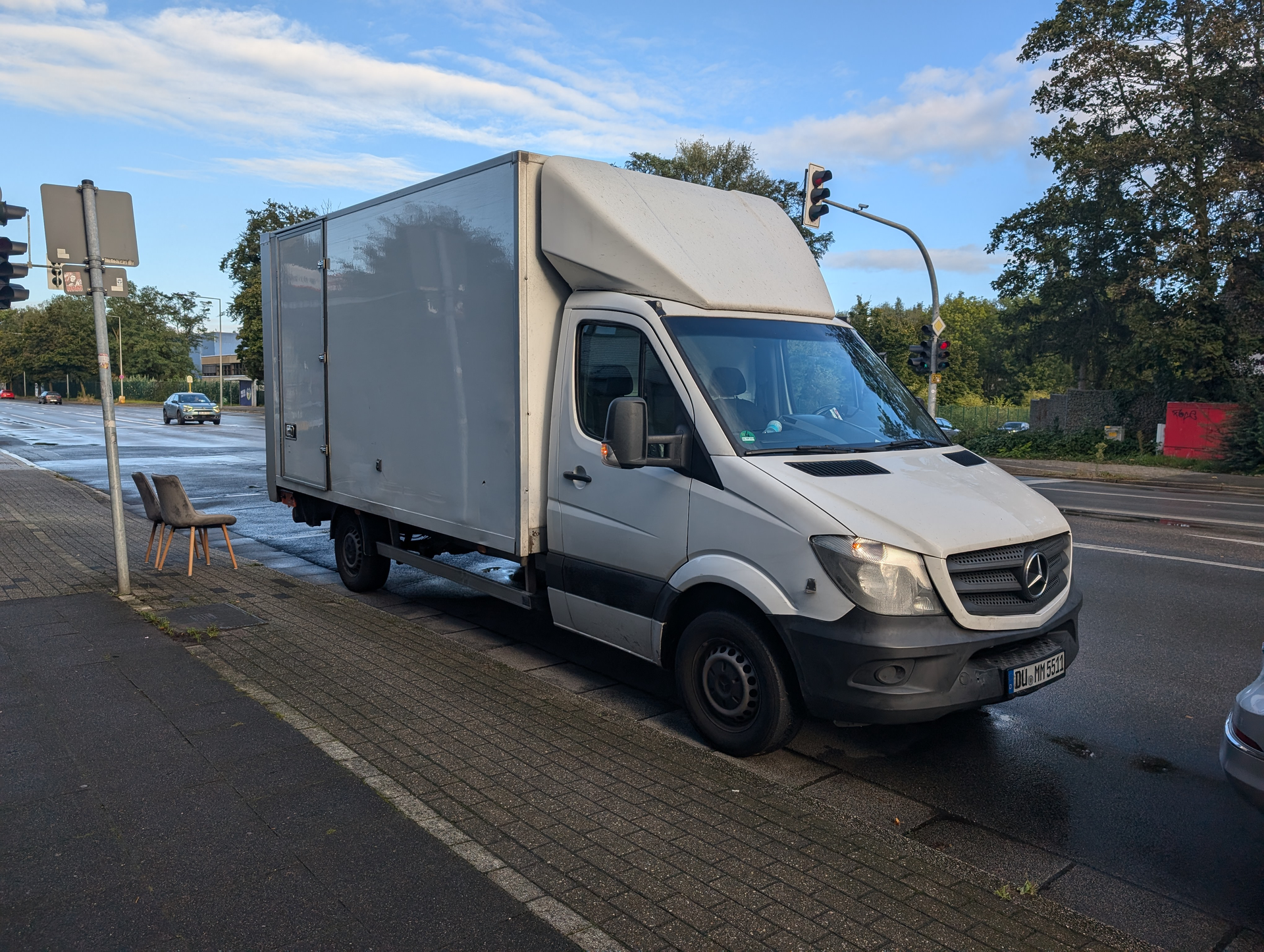 Weißer Kastenwagen, der am Straßenrand neben einer Bank, einer Ampel und einem Zebrastreifen geparkt ist.