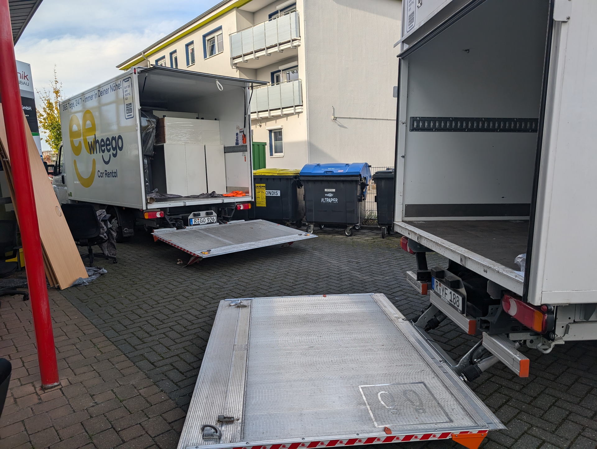 Zwei Lieferwagen mit offenen Hebebühnen; einer lädt ein großes Gerät auf einem gepflasterten Bereich in der Nähe von Gebäuden und Mülltonnen.