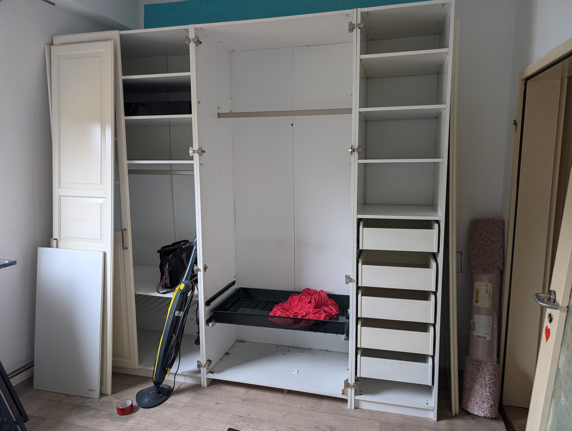 Weißer Schrank mit offenen Türen, Regalen, Schubladen und Kleiderstange in einem Zimmer.