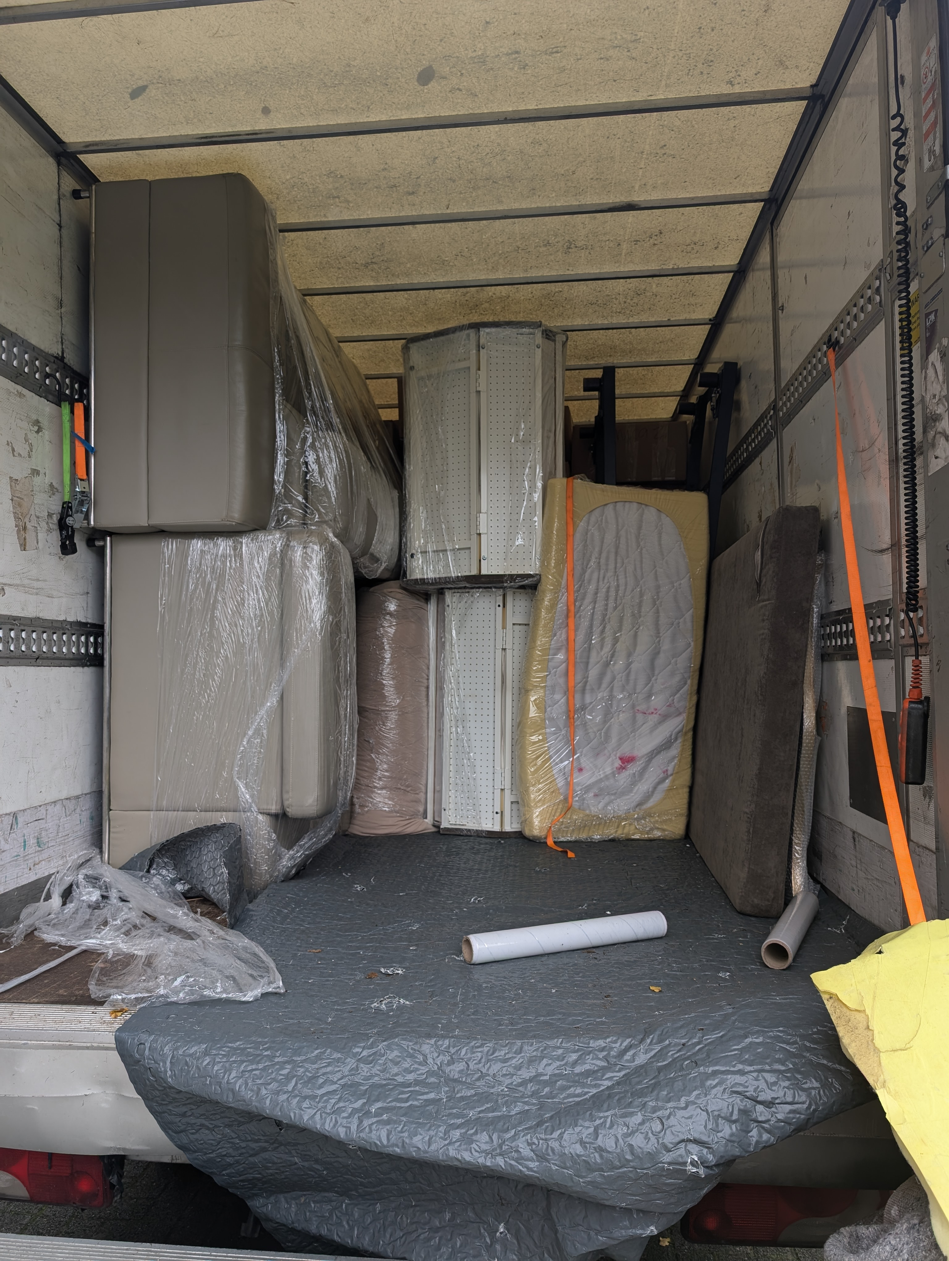 Im Inneren eines Umzugswagens, der mit in Plastik eingewickelten Möbeln beladen ist. Beige und braune Gegenstände sind mit Gurten gesichert.