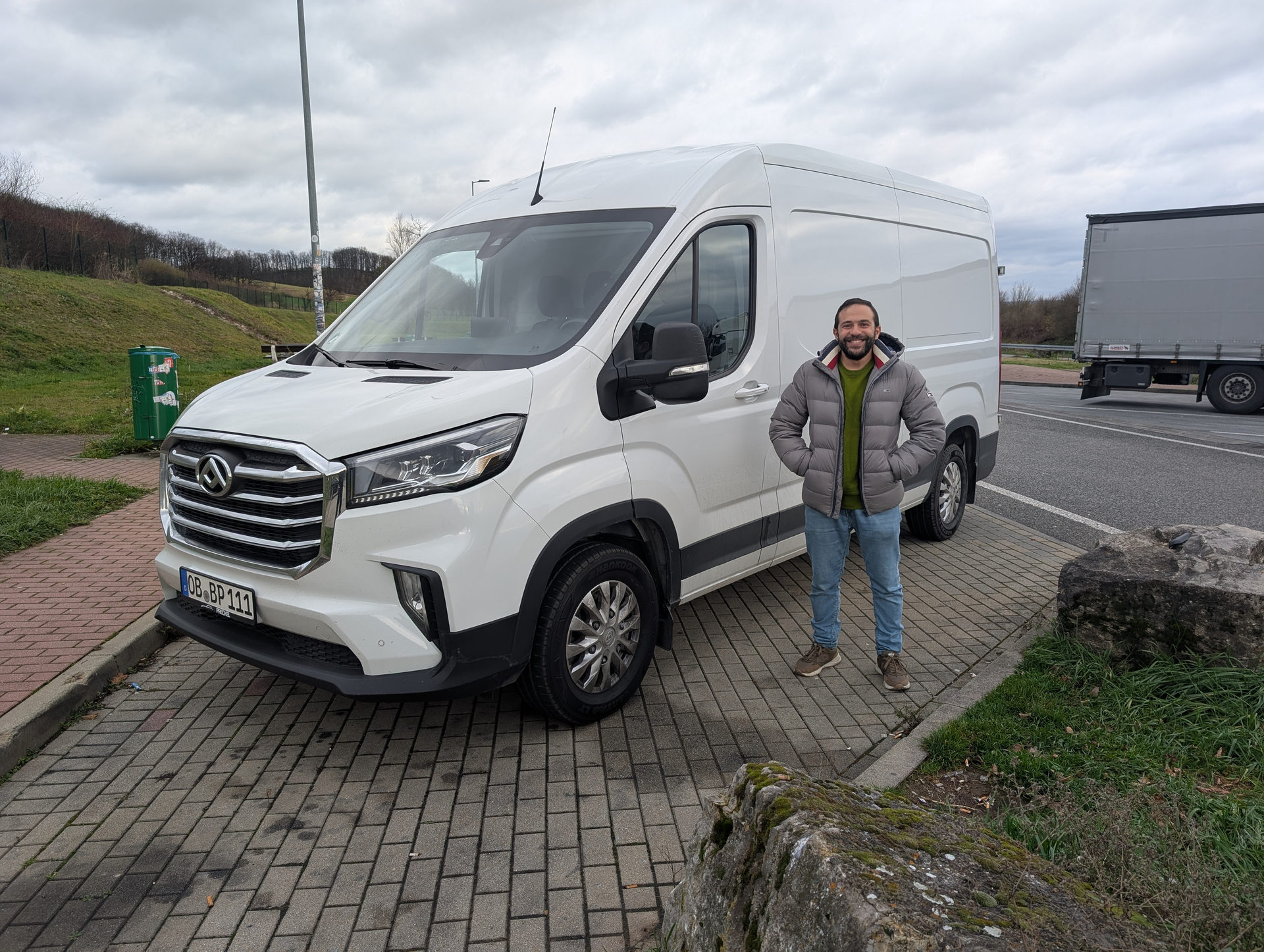 Mann steht neben einem weißen Lieferwagen, der im Freien auf einer gepflasterten Fläche in der Nähe einer Straße geparkt ist.