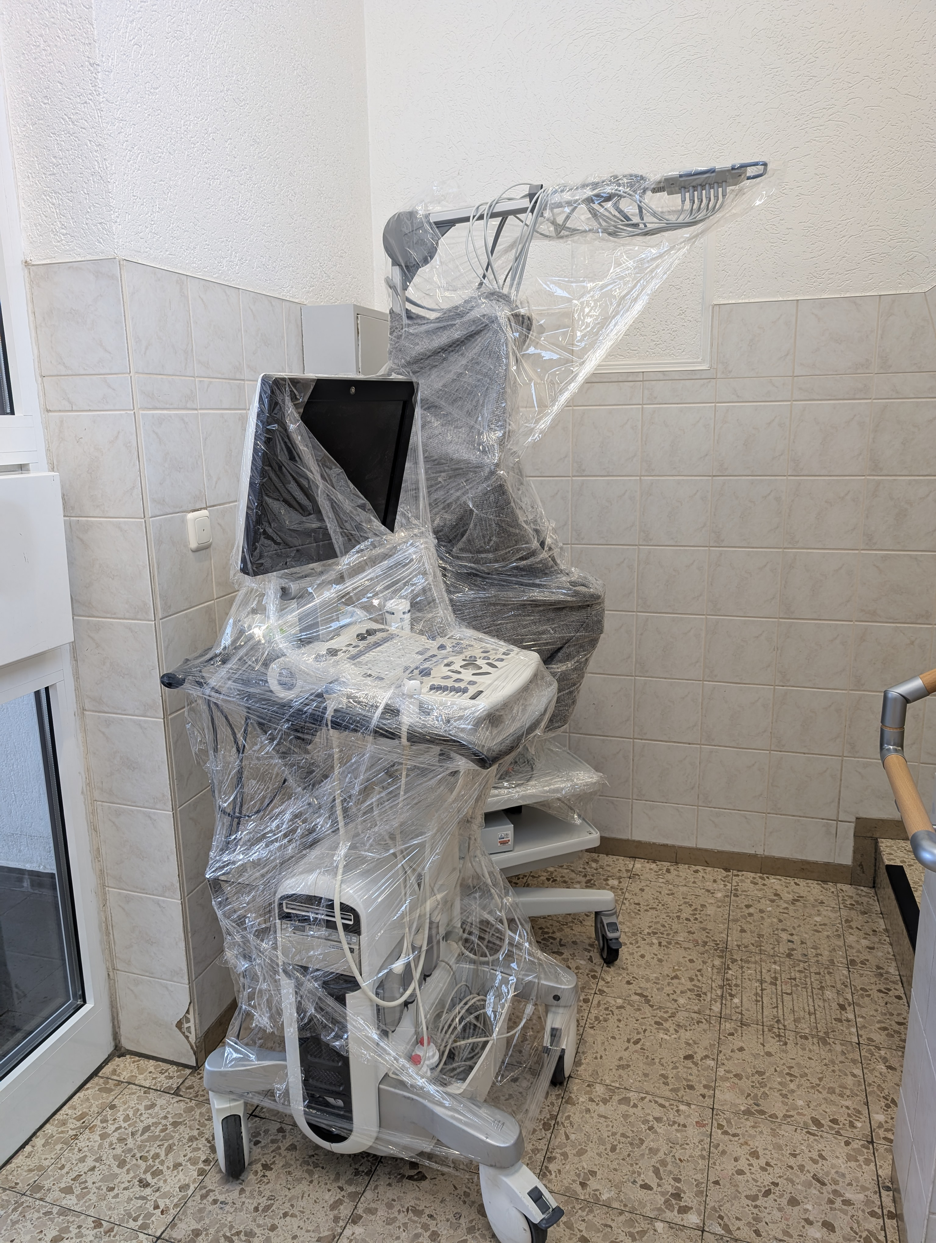 Medizinisches Ultraschallgerät, in Plastik eingewickelt, an einer gefliesten Wand in einer medizinischen Umgebung positioniert.