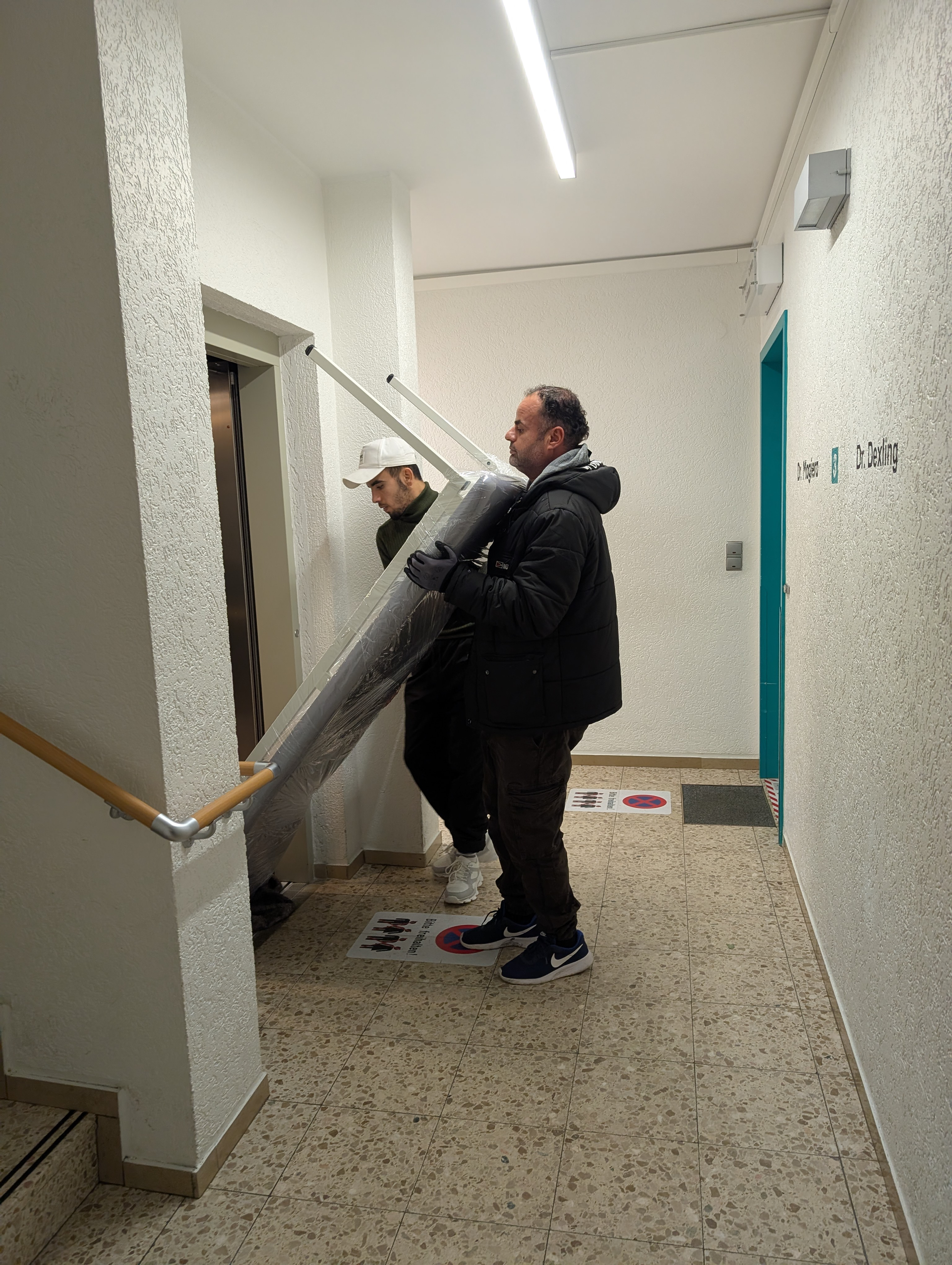 Zwei Männer tragen einen großen, zusammengerollten Gegenstand in einem Flur in Richtung eines Aufzugs.