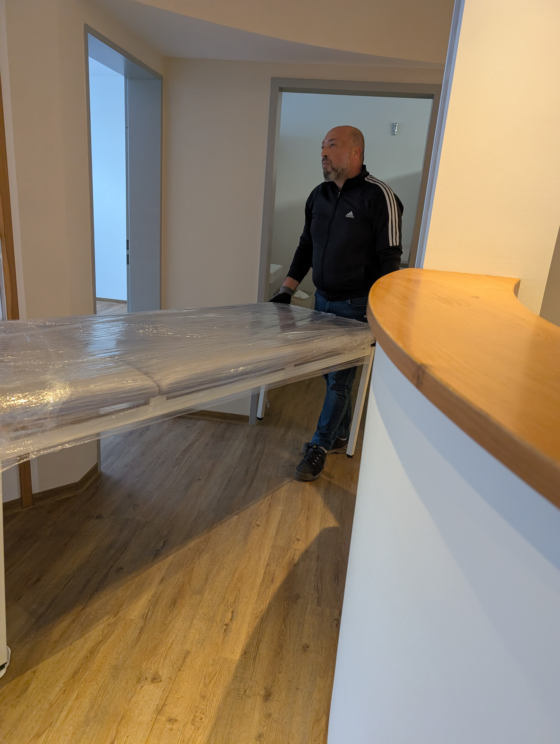 Mann trägt einen verpackten weißen Tisch durch einen Türrahmen, Holzboden, helles Interieur.