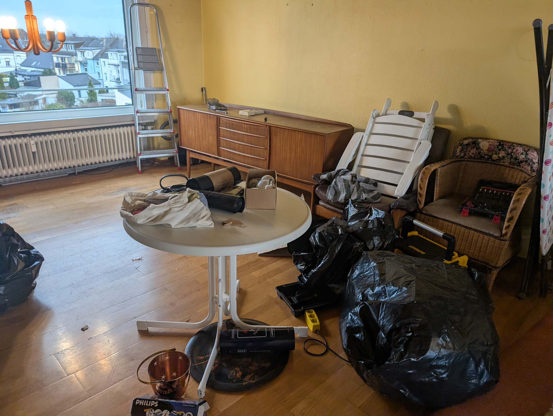 Ein überfülltes Wohnzimmer mit Möbeln, einer Leiter und schwarzen Müllsäcken. Gelbe Wände und ein Blick aus dem Fenster.
