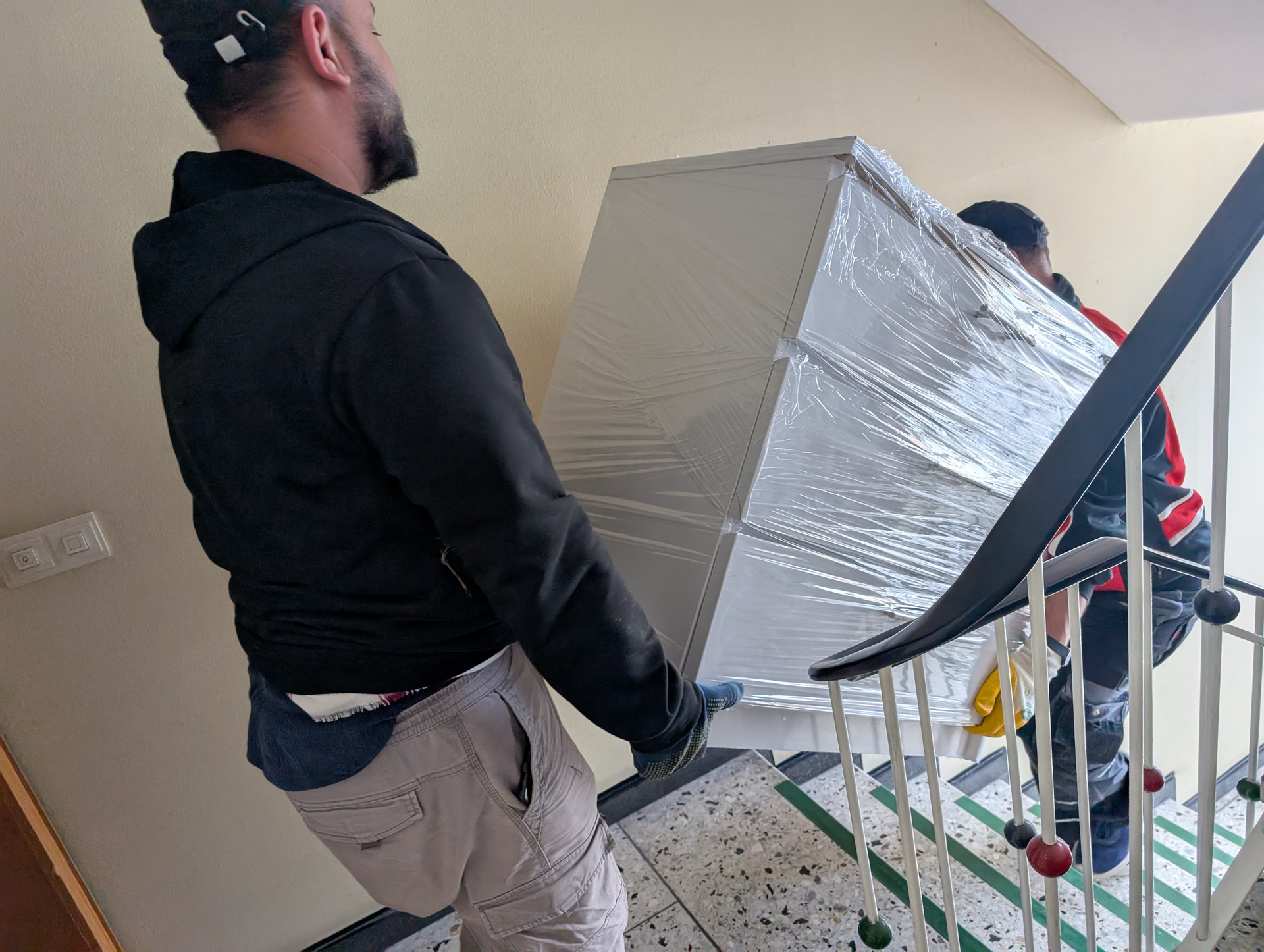 Zwei Möbelpacker tragen einen verpackten, weißen Karton eine Treppe hinauf. Einer trägt eine schwarze Jacke und einen Hut, der andere rot-blaue Kleidung.