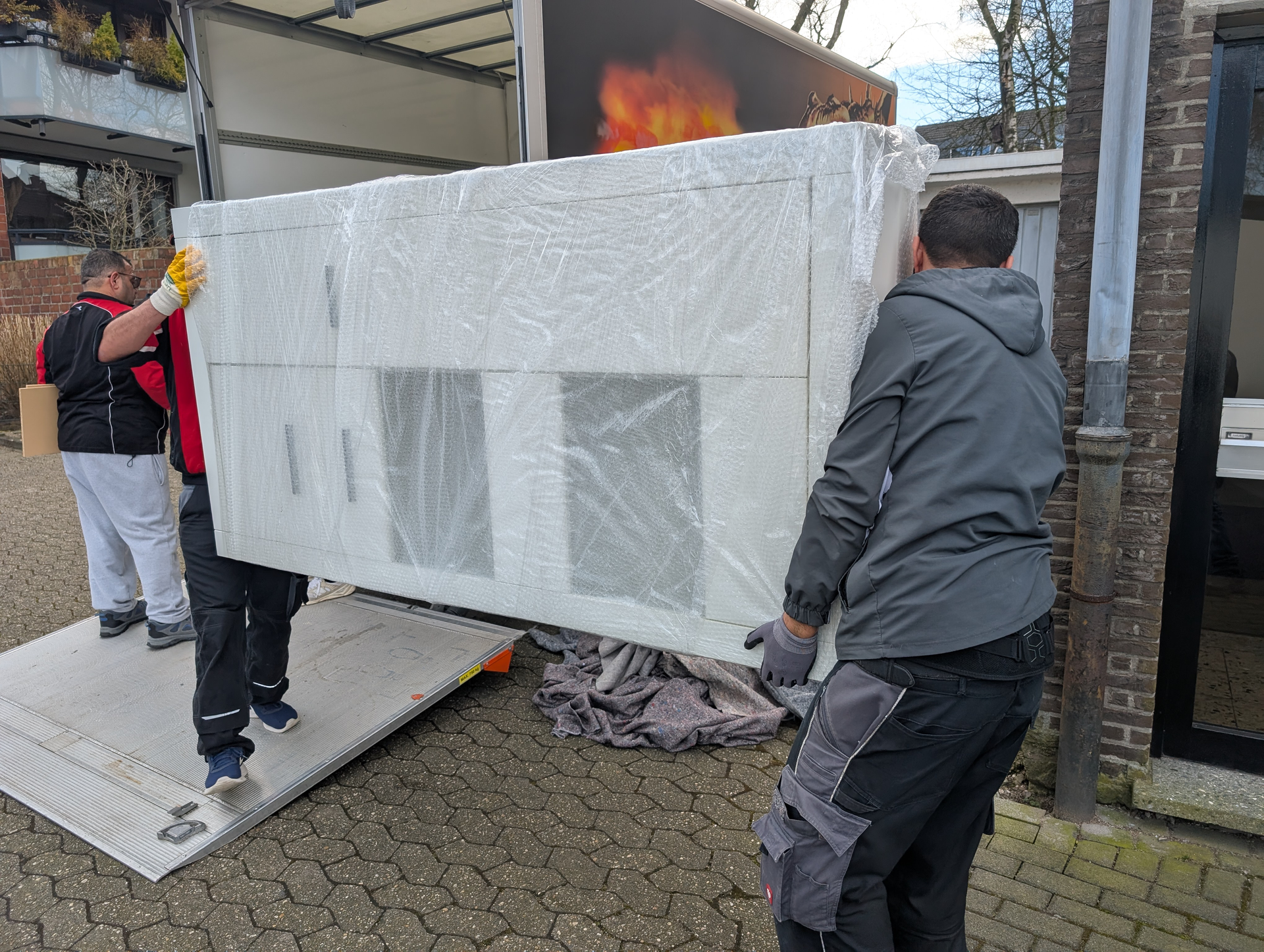 Drei Möbelpacker laden einen großen, weiß verpackten Schrank in einen LKW. Draußen, bei Tageslicht.
