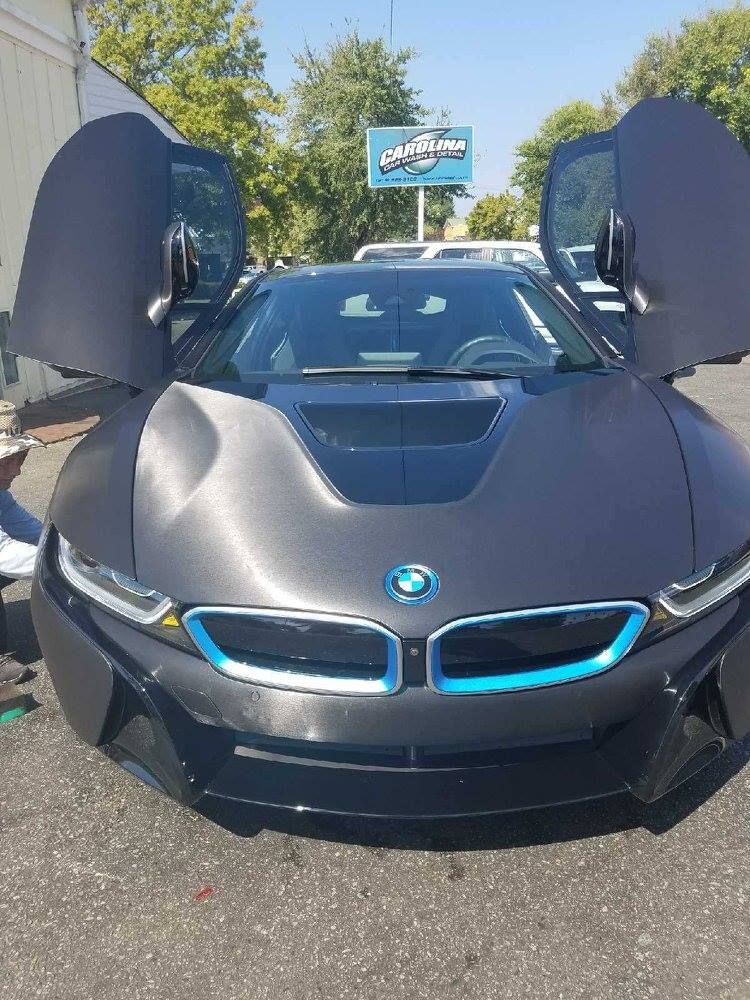 Gray BMW i8 with butterfly doors open, parked in front of a shop.
