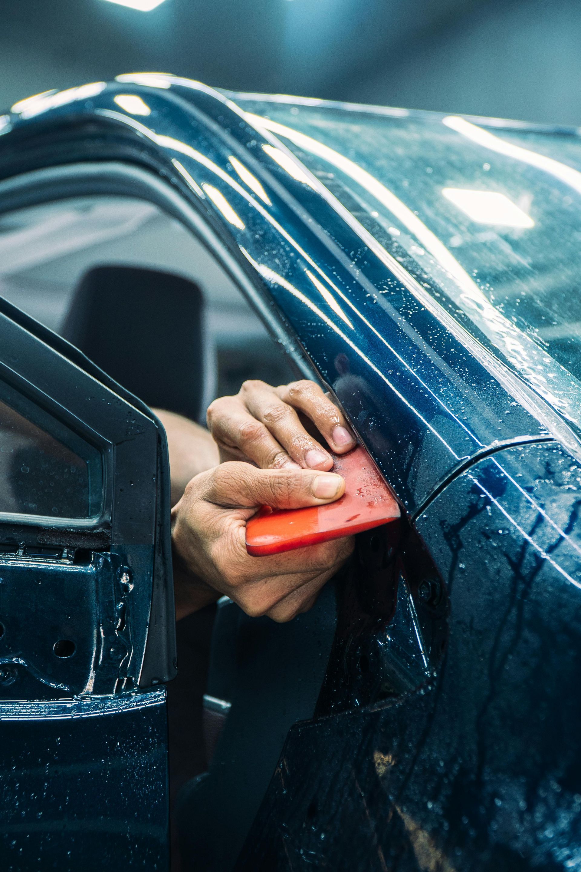 Hand holding a red sponge to wash a blue car window with soap and water running down the side