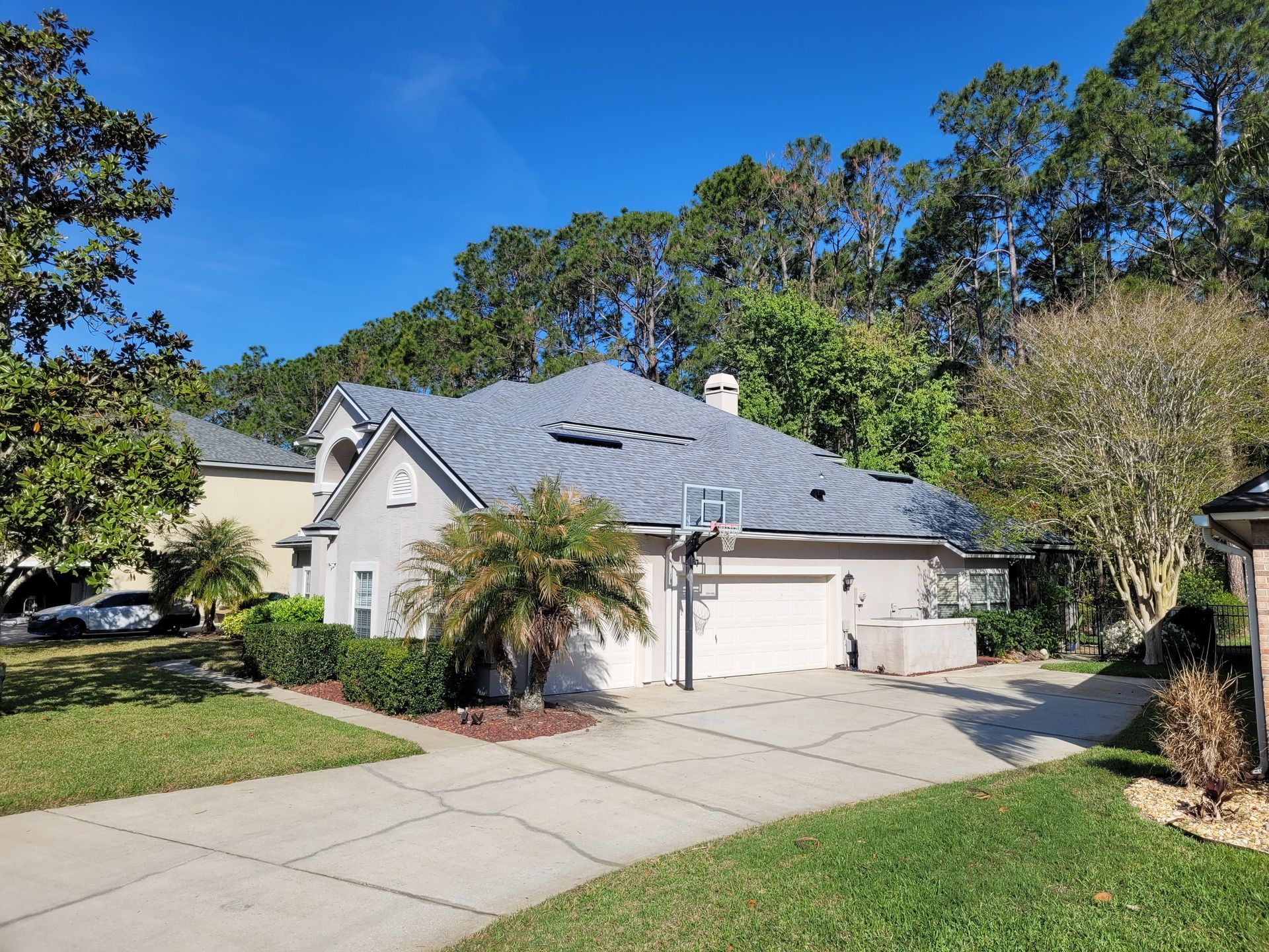 A light stucco house with a gray roof and a basketball hoop in front of a blue sky and trees.
