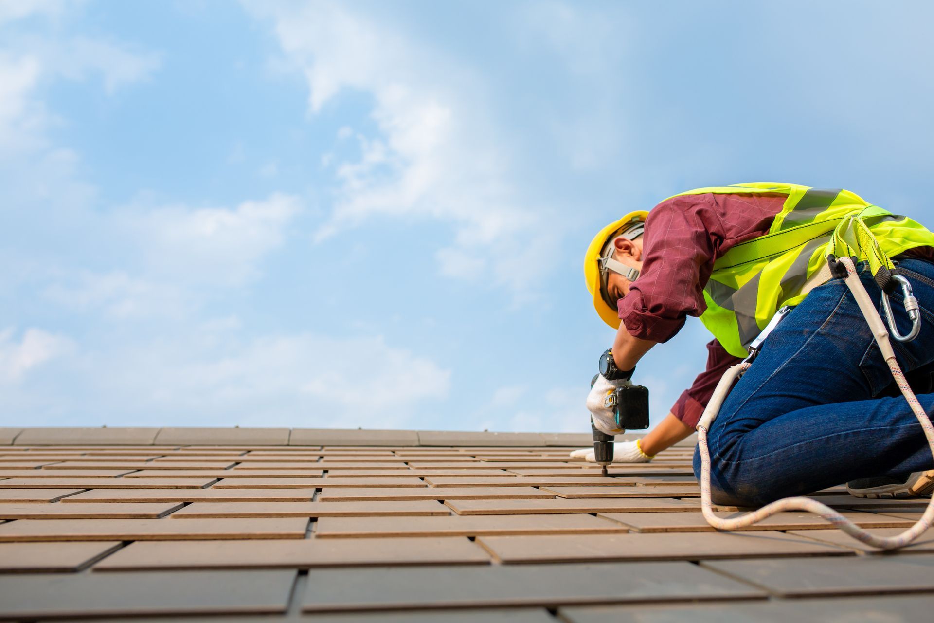 Roofer wearing safety gear, working on a rooftop with a drill against a blue sky.
